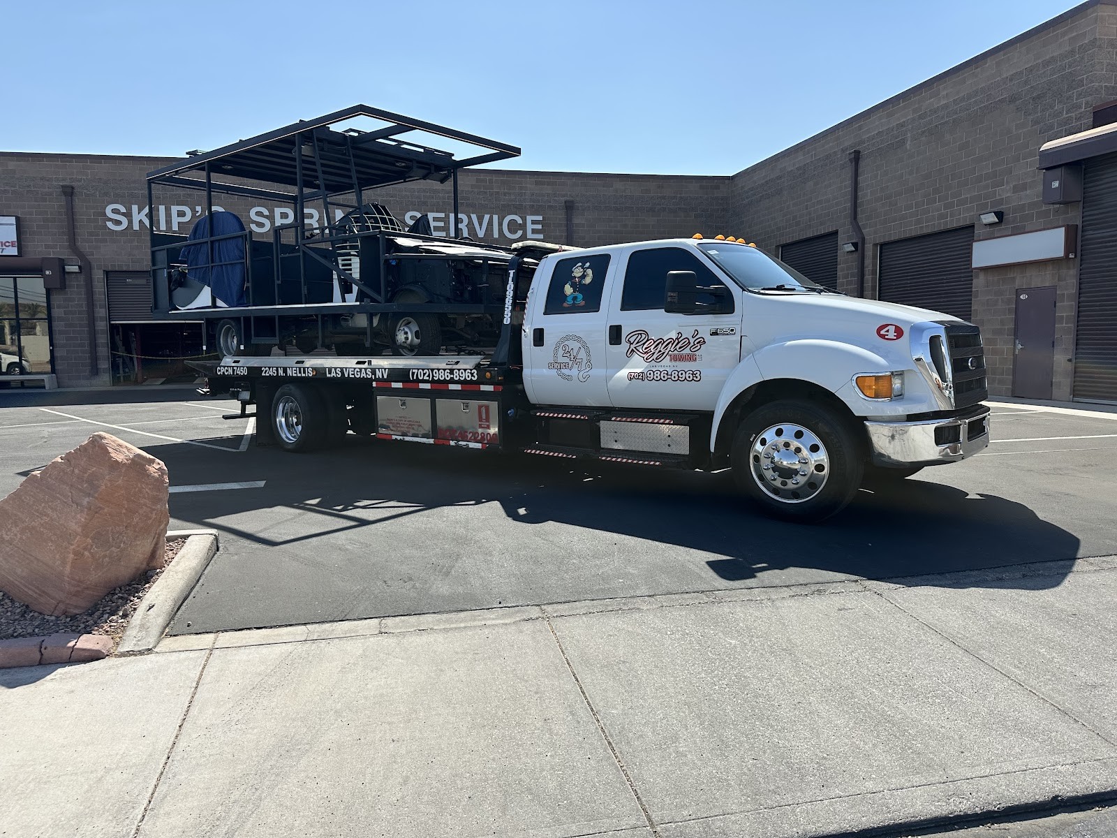 Reggie's Towing flatbed truck parked outside Skip's Spring Service hauling equipment